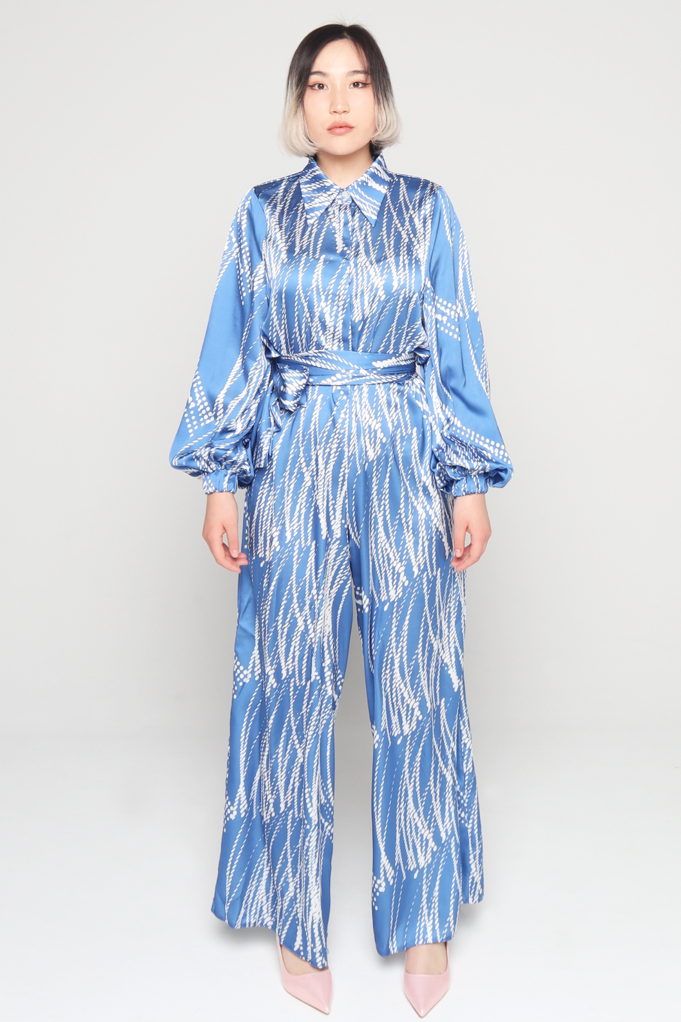 Woman wearing Blue and white collared hidden buttons side pockets elasticated balloon sleeves beautifully patterned satin Jumpsuit falls loosely through the legs with detachable belt A perfect Tznius style summer outfit for women